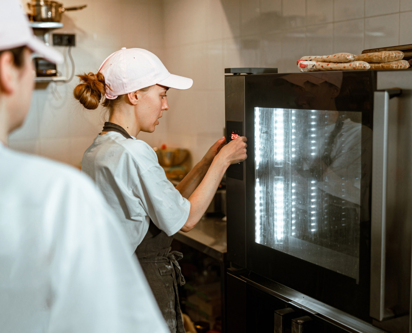 Woman in cap presses keys on control panel of oven with colleague in workshop